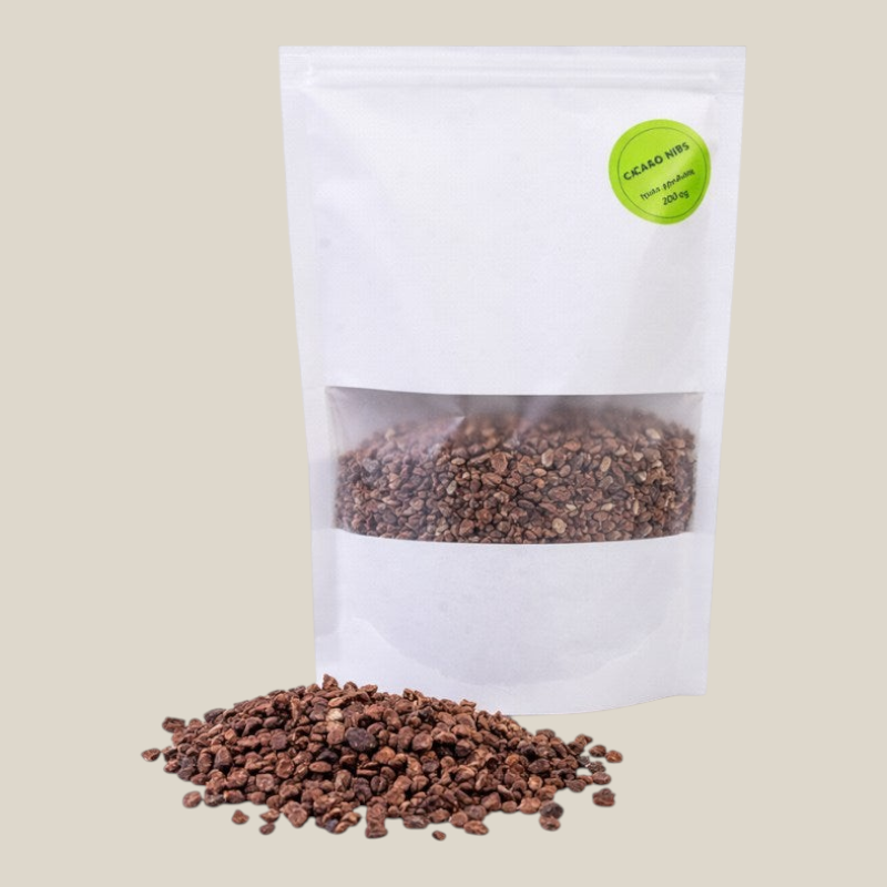 TuanaShop BV cacao nibs in een witte stazak met venster, getoond met een hoopje rauwe cacaostukjes op de voorgrond.
